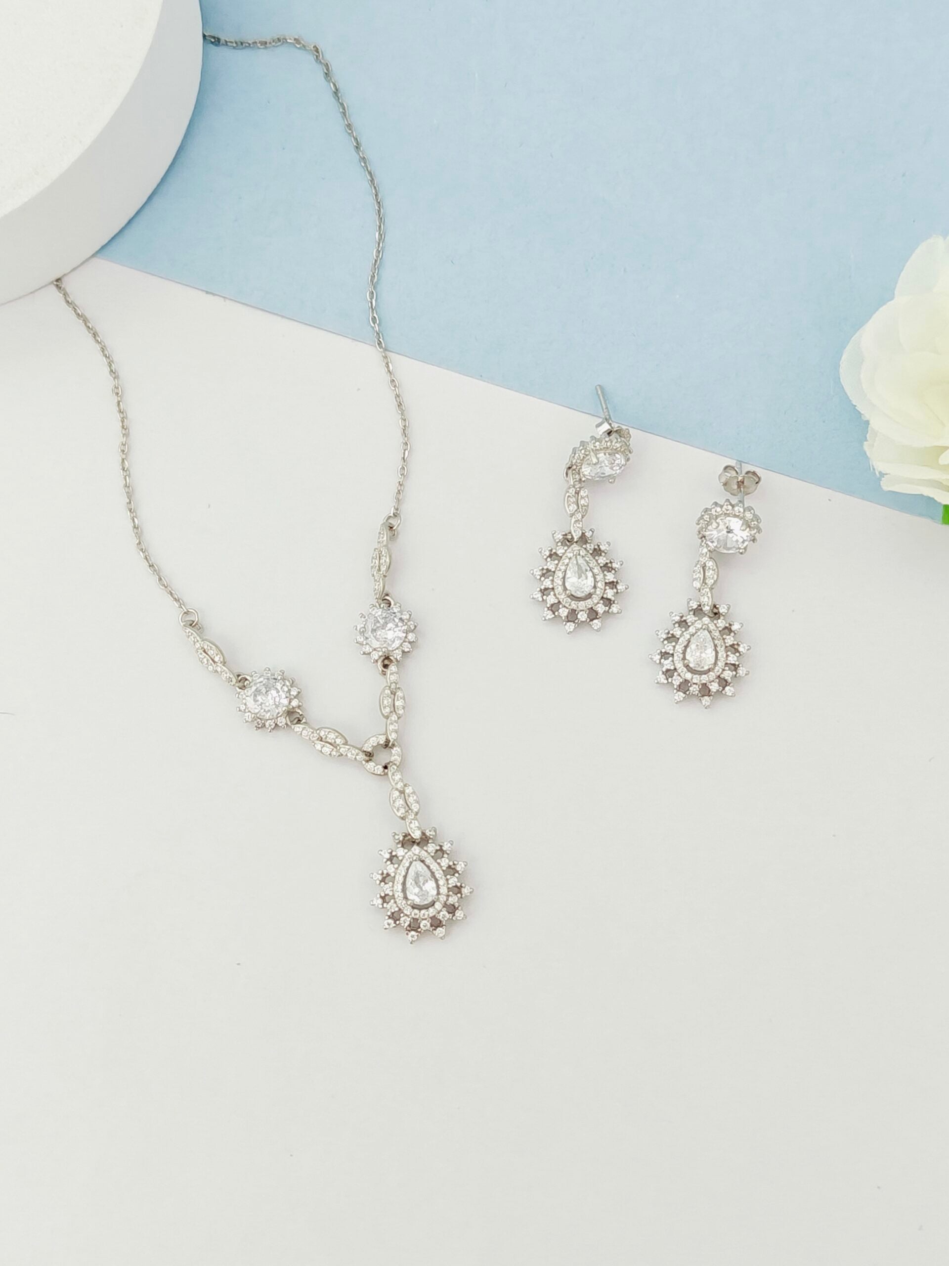 Silver necklace and earrings set on a white surface with a light blue background